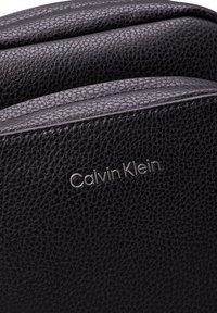 Czarna skórzana torba o teksturowanej powierzchni, z gładkim wykończeniem i srebrnym logo "Calvin Klein" na przodzie.
