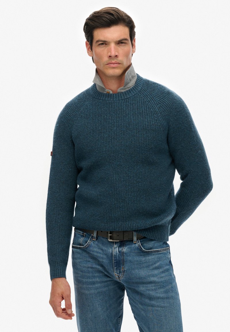 Dunkelblauer Strickpullover mit gerippter Textur, Rundhalsausschnitt und Raglanärmeln, kombiniert mit hellblauen Jeans.