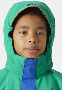 Zielona wodoodporna kurtka z niebieskim zamkiem, wysokim kołnierzem i kapturem. Wnętrze kaptura wykończone jest na pomarańczowo. Posiada logo na górze.