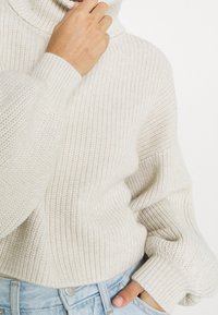 Person som bär en krämfärgad grovstickad polotröja och ljusblå jeans, med en hand nära halsen och den andra i fickan.