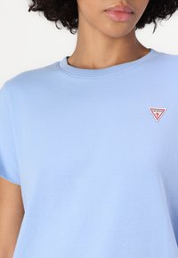 Tricou din bumbac de culoare albastru deschis, cu guler rotund și mâneci scurte, având un mic logo Guess triunghiular roșu și alb pe partea stângă a pieptului.