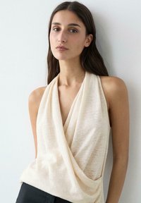 Junge Frau mit langem braunem Haar, die ein ärmelloses, beiges, drapiertes Oberteil trägt und mit neutralem Ausdruck vor einer weißen Wand steht.