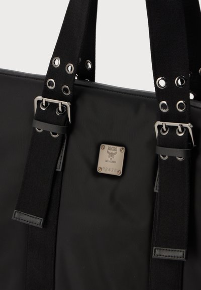 Sac noir avec de larges sangles en tissu comprenant des œillets et boucles métalliques, ainsi qu'une étiquette de marque métallique MCM München avec numéro de série.