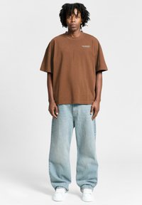 Brown oversized cotton t-shirt with graphic text on chest, paired with light blue baggy denim jeans and white sneakers.