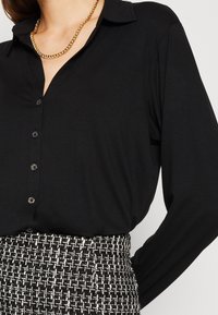Femme portant une chemise noire boutonnée, un collier chaîne doré et une jupe en tweed à motif noir et blanc.