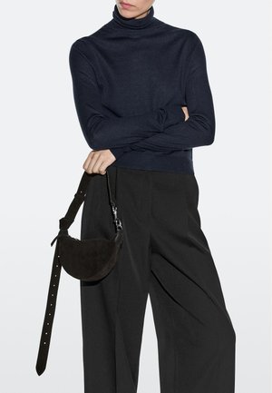 Persona che indossa un dolcevita blu navy e pantaloni neri ampi, che tiene una borsa a mezza luna in suede nero con una tracolla lunga e regolabile.