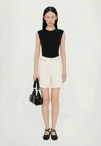 Top negro sin mangas, pantalones cortos crema con bolsillos frontales, sosteniendo un pequeño bolso negro y usando zapatos negros con correas en el tobillo.