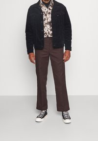 Veste en velours côtelé noire sur une chemise à motifs bruns, association avec un pantalon marron et des baskets noires avec des semelles et des lacets blancs.