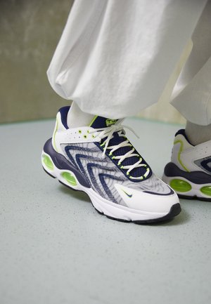 Zapatillas blancas y azul marino con detalles en verde neón, usadas con calcetines blancos y pantalones blancos holgados sobre un suelo gris claro.
