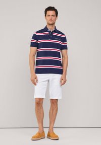 Camiseta polo azul marino con rayas horizontales rojas y blancas, de manga corta y con cuello. Combinada con pantalones cortos blancos y zapatos marrones de tipo slip-on.