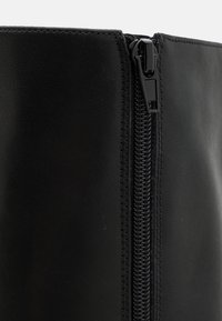 Crne kožne čizme s prednjim zatvaranjem na patentni zatvarač, glatkom teksturom i elegantnim cilindričnim oblikom. Suptilni detalji šivanja duž šavova.