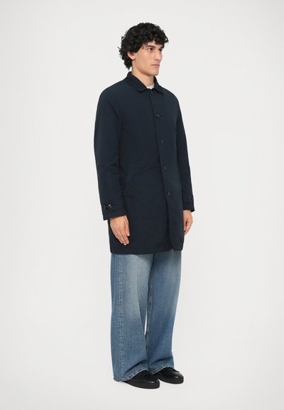 Mørkeblå frakke med krave, frontknapper og sidelommer, parret med brede denimjeans og sorte sko.