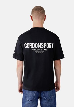 Mann mit kurzen Haaren, der ein schwarzes T-Shirt mit weißem "CORDONSPORT ATHLETICS 1986" Text auf dem Rücken und blaue Jeans trägt, von hinten gesehen.