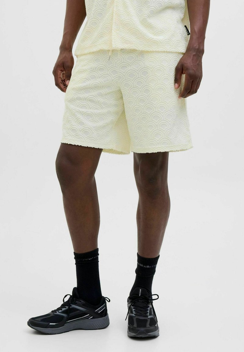 Pantalones cortos amarillos claros con un patrón de olas texturizado y bordes deshilachados, combinados con zapatillas deportivas negras y calcetines hasta el tobillo.