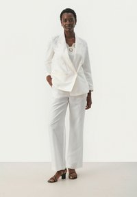 Hvid linned blazer med dobbeltradede knapper og brede bukser. Gennemsigtig top med blomsterhæklede mønstre. Sorte strophæle.