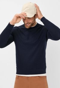 Navy Strickpullover mit Rundhalsausschnitt und langen Ärmeln, kombiniert mit einer hellbeigen Mütze. Die Textur wirkt weich und enganliegend.