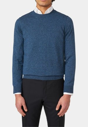 Homme portant un pull bleu à col rond par-dessus une chemise blanche à col et un pantalon noir, debout devant un fond uni.