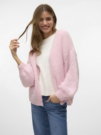 Donna sorridente che indossa un morbido cardigan rosa pastello sopra una camicia bianca e jeans blu, tenendo una ciocca dei suoi lunghi capelli castani.