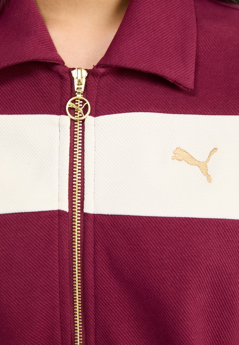 Bordeauxfarbige zip-up Jacke mit weißen Streifen, goldener Reißverschluss und Puma-Logo. Hergestellt aus strukturiertem Stoff mit Kragen.