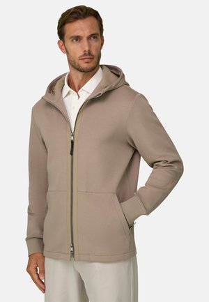 Homme portant un sweat à capuche marron clair zippé sur une chemise blanche à col, une main dans la poche, regardant légèrement sur le côté.
