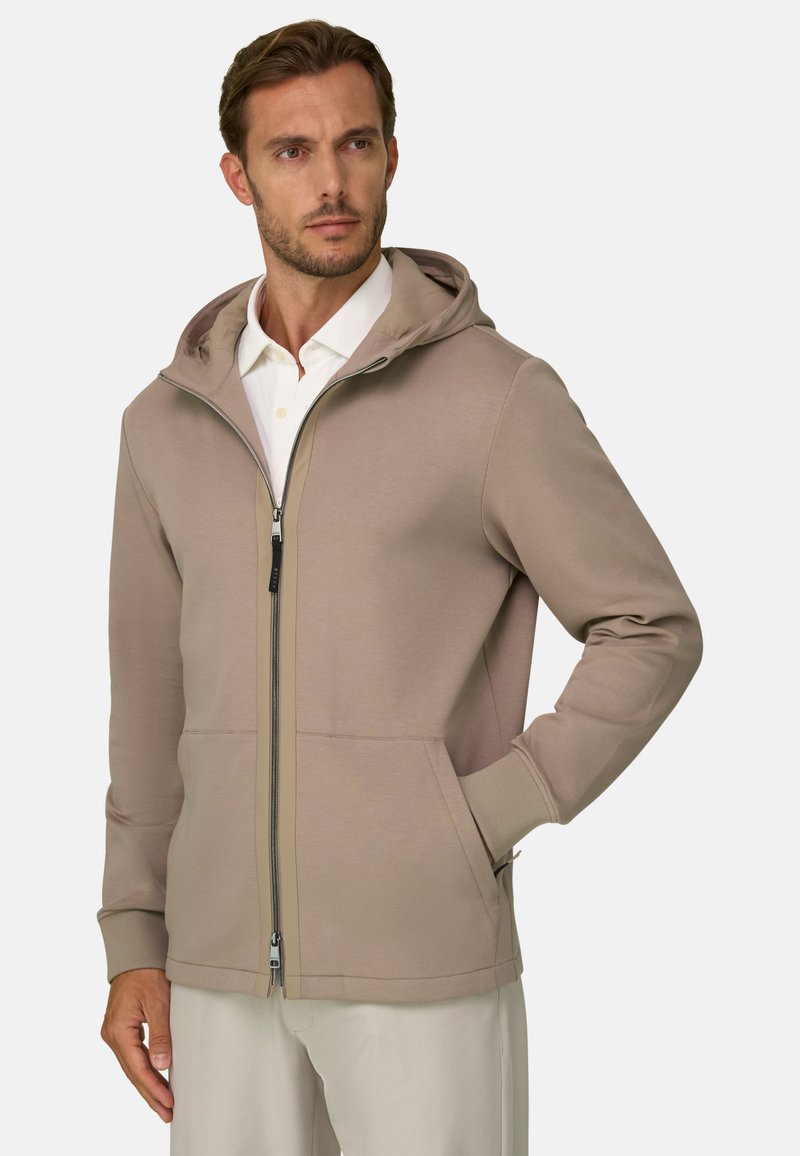 Homme portant un sweat à capuche marron clair zippé sur une chemise blanche à col, une main dans la poche, regardant légèrement sur le côté.