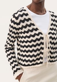 Cardigan tricoté noir et crème avec un motif ondulé, design court, bords festonnés et liens à l'avant, porté sur un haut blanc.