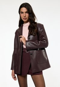 Dunkelburgunderfarbener Leder-Blazer mit einem Knopfverschluss, zwei Fronttaschen und einer entspannten Passform, kombiniert mit einem hellrosa Oberteil und einem gestreiften Rock.