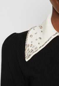 Tissu noir côtelé avec un col blanc orné d'accents argentés et un mélange de strass ronds et rectangulaires.