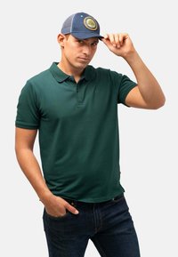 Mörkgrön pikétröja med korta ärmar, tvåknappsslutning och slim fit, kombinerad med en blå keps som har en cirkulär trädlogotyp.