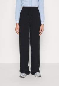 Person som bär svarta vida byxor med dragsko, en ljusblå långärmad skjorta med knappar och silvriga sneakers, stående mot en vit bakgrund.