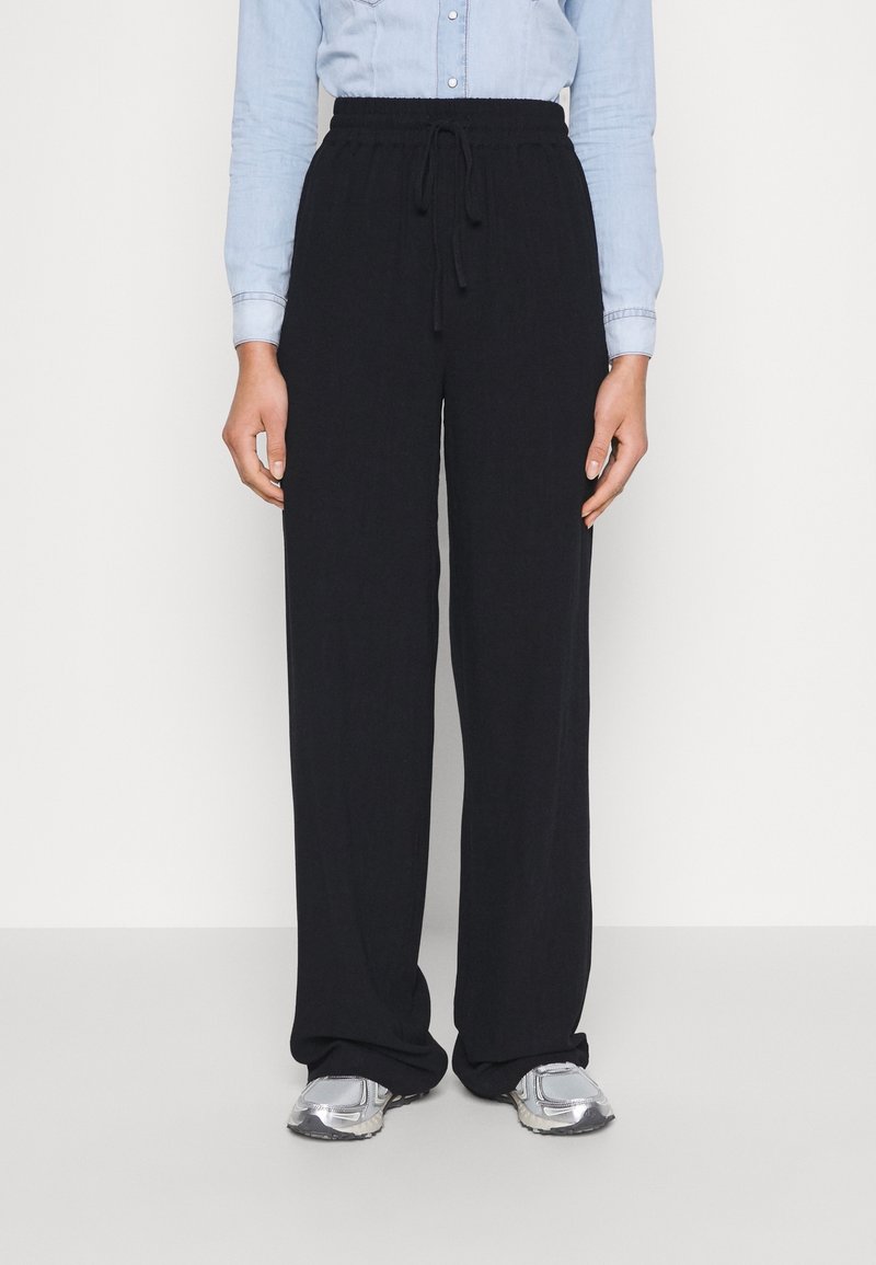 Person som bär svarta vida byxor med dragsko, en ljusblå långärmad skjorta med knappar och silvriga sneakers, stående mot en vit bakgrund.