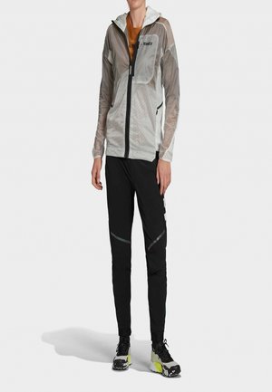 Person trägt transparente weiße Kapuzenjacke, orangefarbenes Hemd, schwarze Hose mit reflektierenden Akzenten und schwarze, weiße und gelbe Sneakers.