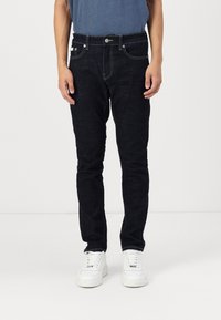 Dark blue jeans made of denim, slim fit, five pockets, with light stitching and metal button hardware, paired with white sneakers.
