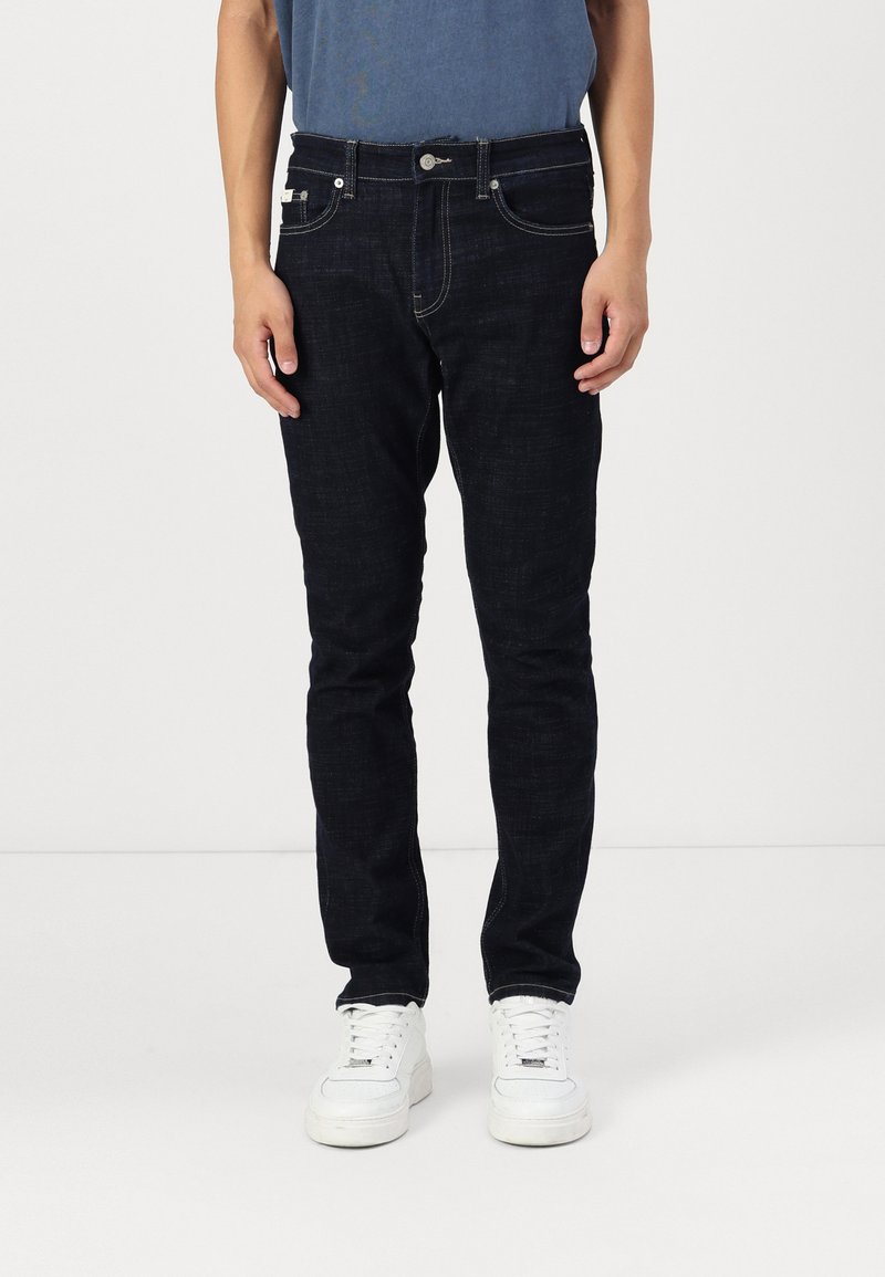 Dark blue jeans made of denim, slim fit, five pockets, with light stitching and metal button hardware, paired with white sneakers.