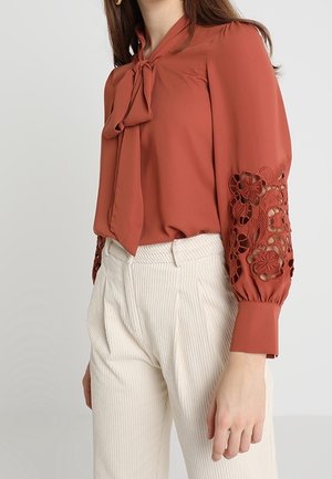 Femme portant un chemisier couleur rouille avec des empiècements en dentelle florale sur les manches et un col noué en forme de nœud, associé à un pantalon taille haute à fines rayures blanches.