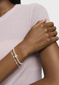 Dos pulseras en una muñeca: una presenta acentos plateados y detalles de perlas, la otra es una cadena de pequeñas perlas, sobre una blusa rosa.