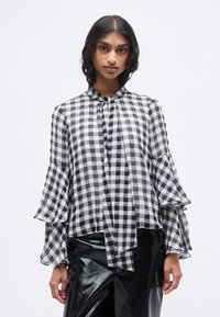 Μπλούζα με καρό μοτίβο σε μαύρο και άσπρο, με μανίκια με βολάν, ψηλό γιακά και λεπτομέρεια με κορδόνι στον λαιμό, κατασκευασμένη από ελαφρύ, διάφανο ύφασμα.