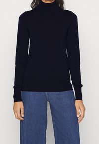 Tummansininen pooloneule sileästä neuloskankaasta, jossa on pitkät hihat ja vartalonmyötäinen malli, yhdistetty vaaleansinisiin leveälahkeisiin farkkuihin.