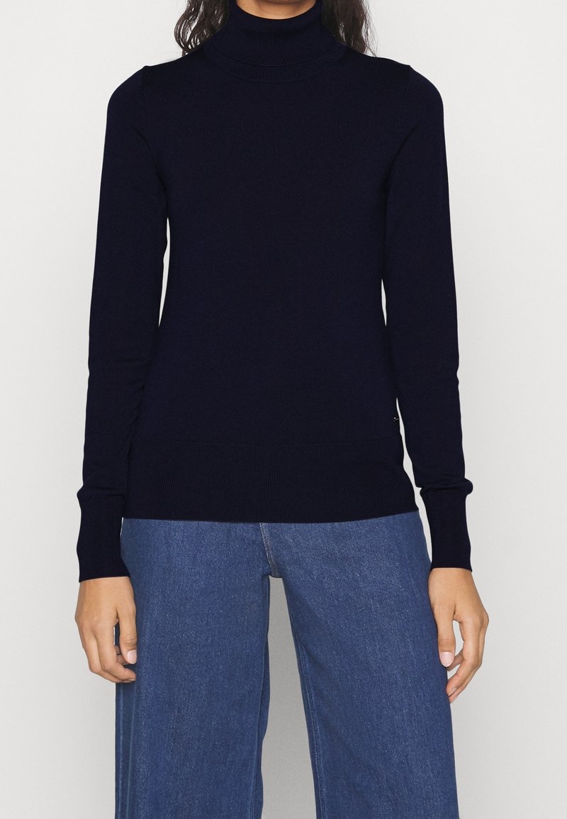 Tummansininen pooloneule sileästä neuloskankaasta, jossa on pitkät hihat ja vartalonmyötäinen malli, yhdistetty vaaleansinisiin leveälahkeisiin farkkuihin.