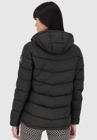 Donna con lunghi capelli scuri che indossa una giacca a vento nera con cappuccio e pantaloni a strisce bianchi e neri, voltata di spalle contro uno sfondo uniforme.