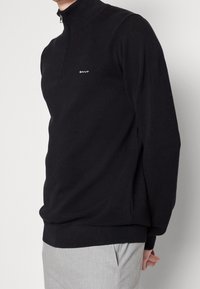 Zwarte pullover met ritssluiting en een gestructureerd gebreid patroon. Heeft een slim fit en een klein wit logo op de borst. Zachte stof.