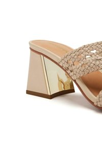 Sandaña de tacón en bloque beige con tira trenzada, tacón con detalles dorados, parte superior suave y plantilla texturizada para mayor agarre.