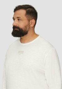 Weißes Langarm-T-Shirt mit rundem Halsausschnitt und einem kleinen beigen Logo-Tag auf der Brust. Hergestellt aus weichem, strukturiertem Stoff.