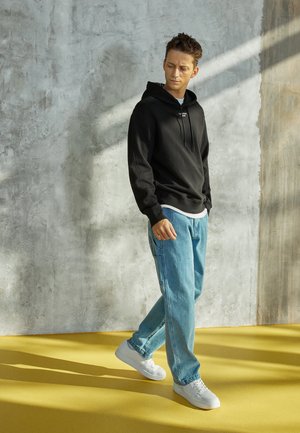 Felpa nera con cappuccio e lacci, indossata sopra una camicia bianca; jeans di denim azzurro chiaro; scarpe da ginnastica bianche; in piedi su un pavimento giallo.