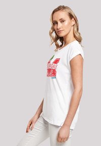 Weißes Kurzarm-T-Shirt aus Baumwolle mit einem bunten Grafikdruck, der Text und Symbole zeigt, kombiniert mit hellen Hosen.