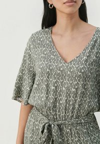 Zelena obleka s potiskom, z V-izrezom, kratkimi rokavi in zavezanim pasom. Tkanina ima kontrastni abstraktni vzorec v beli barvi.