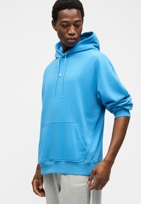 Mann iført en blå hettegenser med forlomme og snorer, kombinert med grå Calvin Klein joggebukser mot en ensfarget lys bakgrunn.