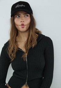 Black ribbed zip-up top with a high collar; worn with a black cap featuring "London Chill Club" in white embroidery. Model makes a playful expression.