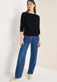 Maglione nero a maglia strutturata con maniche lunghe, abbinato a jeans blu a gamba larga e sneakers beige, in piedi contro uno sfondo chiaro.