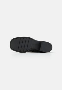 Suela de zapatilla negra con goma texturizada; presenta ranuras horizontales y un logotipo. La superficie es plana, proporcionando tracción y estabilidad.